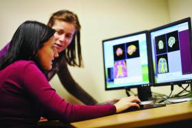 Two women seated at a desk, looking at two computer screens with brain scans on them.
